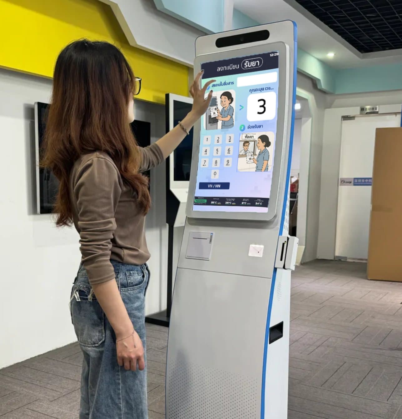 A woman using a medical kiosk with Thai-language interface for hospital registration | หญิงสาวใช้งานตู้บริการทางการแพทย์พร้อมหน้าจอภาษาไทยสำหรับลงทะเบียนในโรงพยาบาล