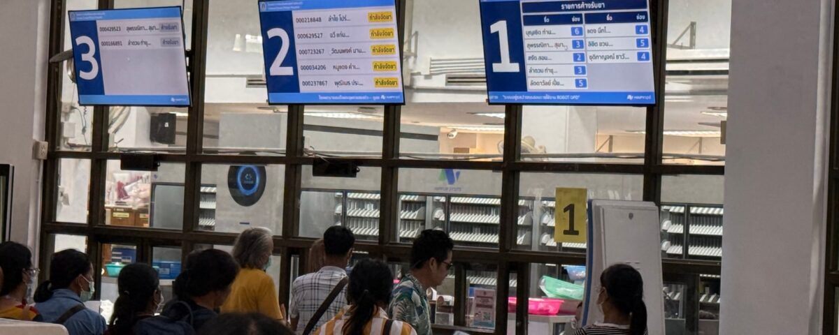 Patients queuing at OPD pharmacy dispensing windows with HAPYopd screens and Robot Pharmacist automated system visible behind glass at Sakaeo Crown Prince Hospital