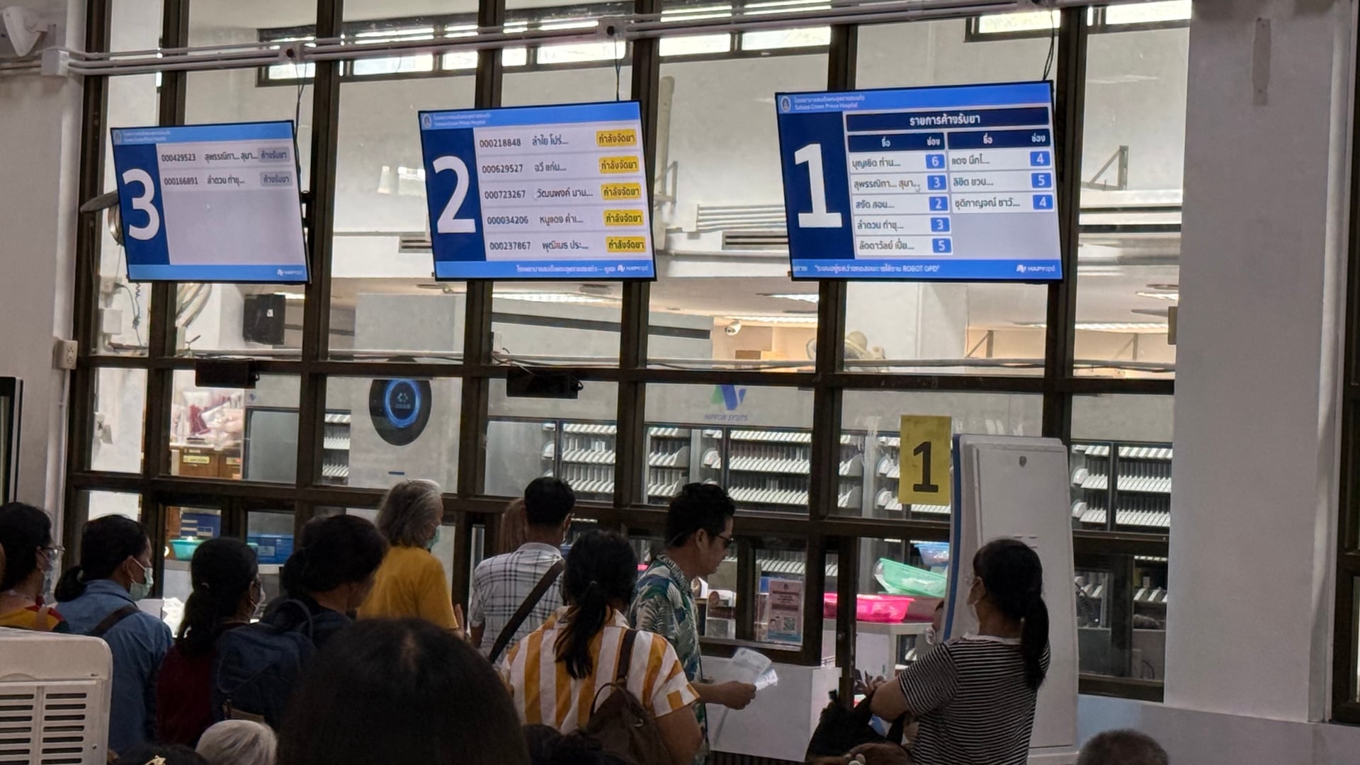 Patients queuing at OPD pharmacy dispensing windows with HAPYopd screens and Robot Pharmacist automated system visible behind glass at Sakaeo Crown Prince Hospital